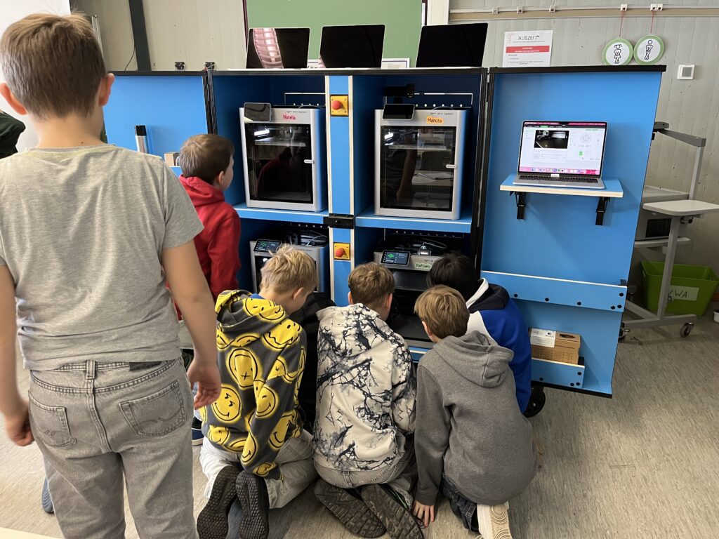 5 Kinder schauen in das Modul mit 4 3D-Druckern. Sie beobachten den Druck.