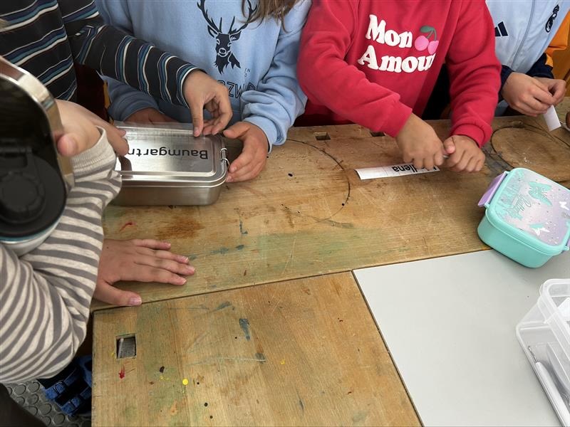 Nahaufnahme von Kinderhänden an einem Holztisch. Ein Kind hält eine silberne Brotdose, auf der der Name „Baumgartn…" mit einem Schneideplotter-Aufkleber aufgebracht wurde. Daneben liegt ein weiterer Namensstreifen „Elena". Im Hintergrund sind weitere Kinder und eine türkisfarbene Brotdose zu sehen.