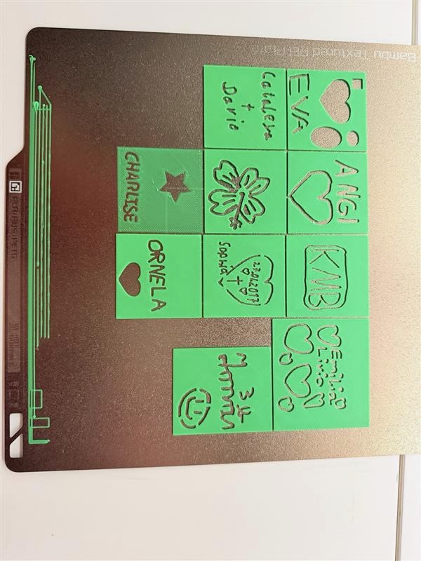 Draufsicht auf eine metallene 3D-Drucker-Druckplatte mit neun grünen, frisch gedruckten quadratischen Schablonen. Jede trägt handschriftlich wirkende Gravuren mit Namen und Motiven: Ornela mit Herz, Charise mit Stern, Eva mit Herz, Angi mit Herz, KMB, Cataleja und Derio, eine Blume, ein Datum sowie weitere persönliche Botschaften.