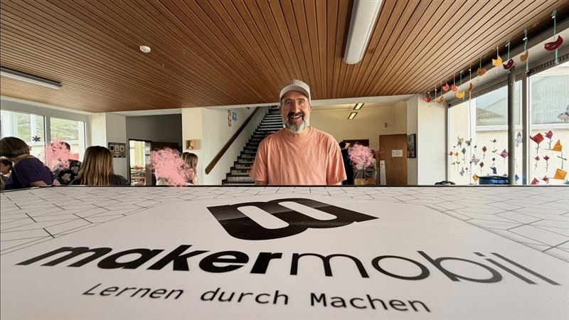 Ein lächelnder Mann mit weißer Cap und lachsfarbenem T-Shirt steht hinter einem weißen Makermobil-Rollcontainer, auf dem das Makermobil-Logo und der Schriftzug „Lernen durch Machen" zu lesen sind. Im Hintergrund ist ein helles Schulfoyer mit Treppe und bunten Fensterdekoration zu sehen. Am linken Rand sitzen Kinder an Tischen.
