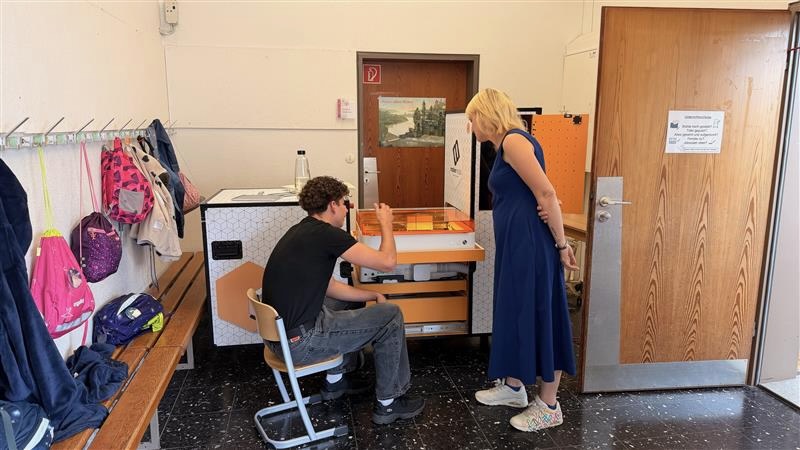 Ein Jugendlicher sitzt vor dem geöffneten orangefarbenen Lasercutter-Modul des Makermobils und bedient das Gerät. Eine erwachsene Frau im blauen Kleid steht neben ihm und schaut zu. Im Hintergrund hängen Schulranzen und Jacken an Haken, an der Wand ist ein Türschild angebracht.