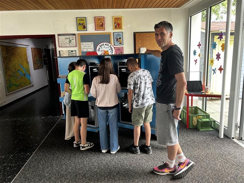 Drei Kinder stehen mit dem Rücken zur Kamera vor dem blauen 3D-Druck-Modul des Makermobils und schauen in die Drucker. Rechts neben ihnen steht ein erwachsener Mann im schwarzen T-Shirt, der in die Kamera schaut. Im Hintergrund ist ein Schulflur mit Landkarte, Plakaten und bunten Blumenmobiles am Fenster zu sehen.