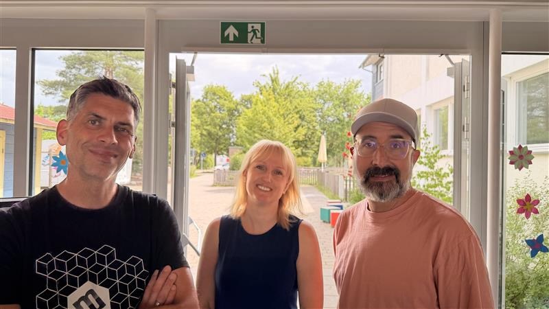 Drei Erwachsene stehen nebeneinander in einem hellen Schulfoyer vor einer gläsernen Ausgangstür. Links ein Mann im schwarzen T-Shirt mit geometrischem Muster, in der Mitte eine Frau mit blonden Haaren und blauem Kleid, rechts ein Mann mit Cap, Brille und lachsfarbenem T-Shirt. Im Hintergrund sind Bäume und ein Schulgebäude zu sehen, an den Wänden bunte Blumendekoration.