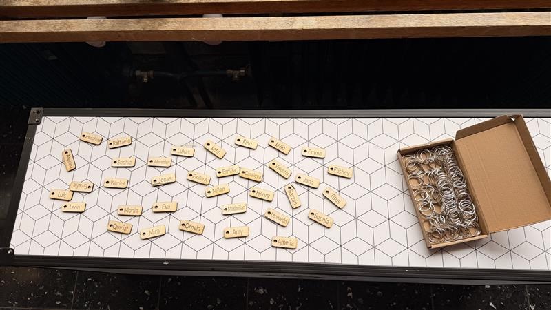 Draufsicht auf einen weißen Makermobil-Rollcontainer mit Hexagonmuster. Darauf liegen zahlreiche kleine rechteckige Holz-Schlüsselanhänger, in die Vornamen eingraviert sind, darunter Josephine, Raffaele, Lukas, Finn, Emma, Leon, Mira, Amelia und viele mehr. Rechts daneben steht eine aufgeklappte Schachtel mit silbernen Schlüsselringen.