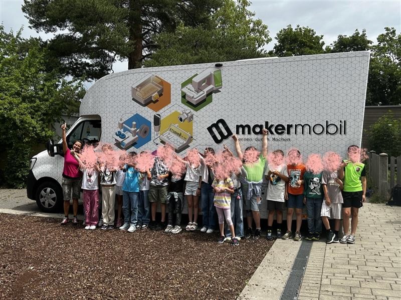 Eine Gruppe von etwa 15 Kindern und zwei Erwachsenen steht vor dem weißen Makermobil-Fahrzeug auf einem Kiesweg. Fast alle blasen gleichzeitig rosa Farbpulver aus ihren Händen in die Luft, sodass ihre Gesichter von einer rosa Pulverwolke verdeckt sind. Auf der Seite des Fahrzeugs sind das Makermobil-Logo sowie Illustrationen der verschiedenen Module zu sehen.