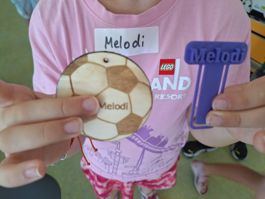 Ein Kind im rosa T-Shirt hält stolz zwei selbst gefertigte Objekte in die Kamera: links ein rundes, lasergraviertes Holzplättchen in Fußballform mit dem eingravierten Namen „Melodi", rechts ein lilafarbenes, 3D-gedrucktes Lesezeichen mit dem aufgedruckten Namen „Melodi". Auf dem T-Shirt des Kindes klebt ein handgeschriebenes Namensschild.

Bild 3 (Heidelberg.jpg):
Das weiße Makermobil-Fahrzeug steht auf einem sonnigen Schulhof vor einem modernen Schulgebäude. Auf der großen Seitenfläche des Fahrzeugs sind das Makermobil-Logo mit dem Schriftzug „Lernen durch Machen" sowie die Logos des Ministeriums für Kultus, Jugend und Sport Baden-Württemberg und des Landesmedienzentrums Baden-Württemberg zu sehen. Im Hintergrund sind Bäume und ein Zaun erkennbar.





