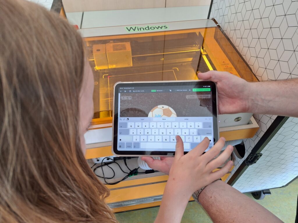 Ein Kind mit langen Haaren tippt auf einem iPad, das vor einem geöffneten orangefarbenen Lasercutter-Modul des Makermobils liegt. Auf dem iPad-Bildschirm ist die Lasercutter-Software zu sehen, in der ein Fußball-Holzplättchen mit dem Namen „Jule" positioniert wird. Eine zweite Person hält das Tablet von der anderen Seite.