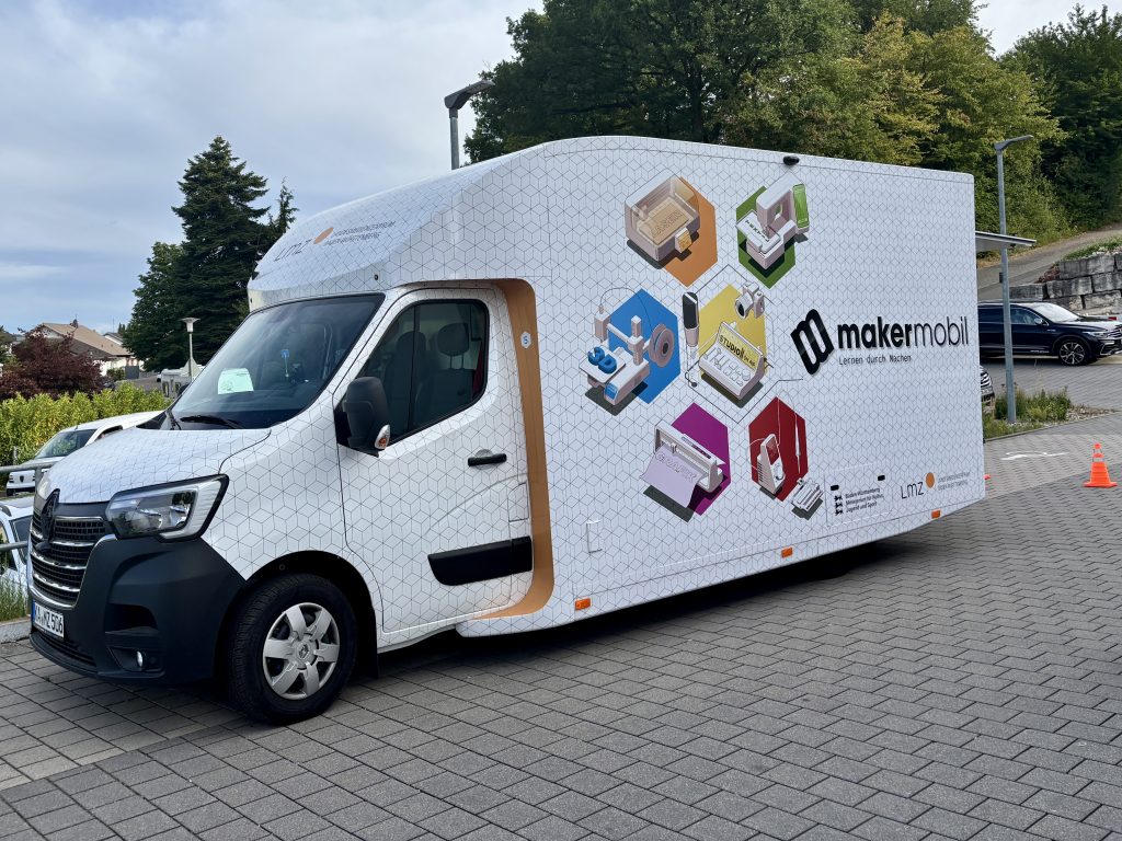 Das Makermobil-Fahrzeug steht auf einem gepflasterten Parkplatz vor einer begrünten Böschung und Bäumen. Die Fahrerseite zeigt das Makermobil-Logo mit dem Schriftzug „Lernen durch Machen" sowie farbige Hexagon-Illustrationen aller sechs Module: 3D-Druck, Grafik, Laser, Mechatronik, Studio und Textil. Unten rechts sind die Logos des Ministeriums für Kultus, Jugend und Sport Baden-Württemberg und des Landesmedienzentrums zu sehen.
