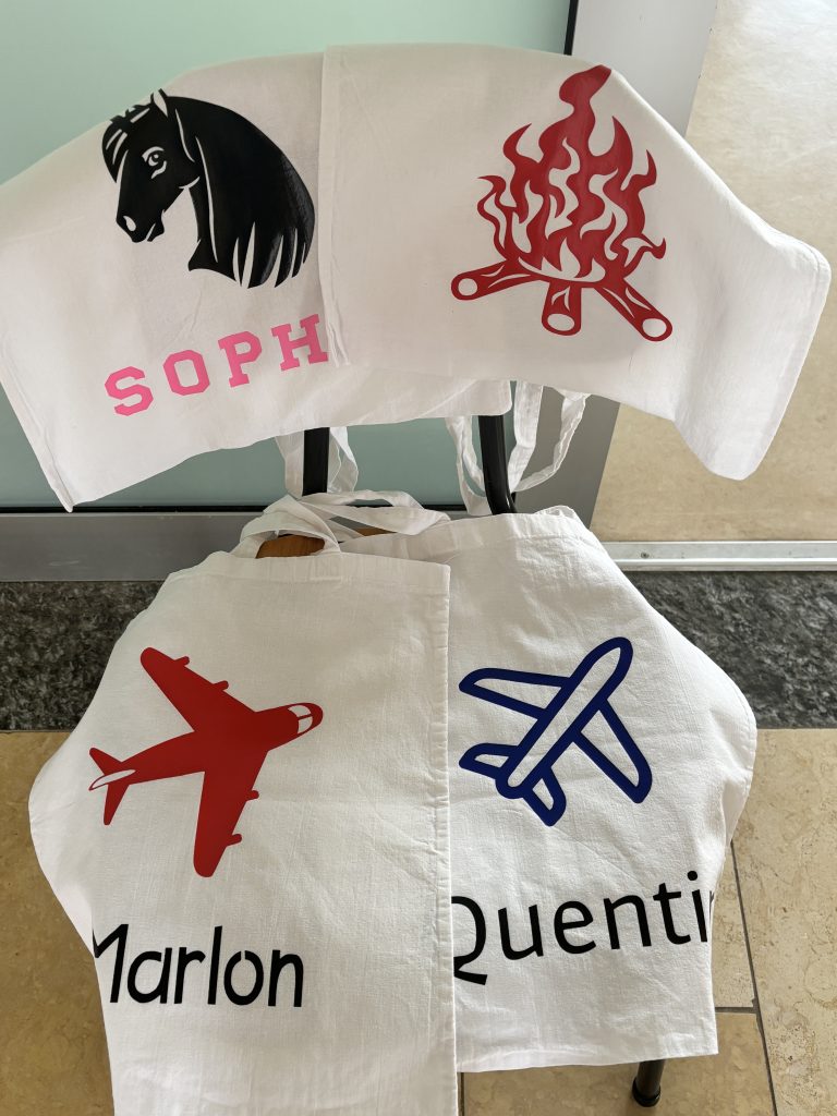 Vier bedruckte Stofftaschen mit individuellen Plotter-Motiven und Namen: Pferd, Lagerfeuer, zwei Flugzeuge.