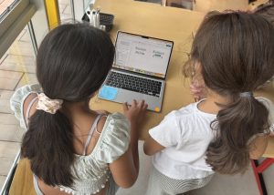 Vogelperspektive auf zwei Mädchen, die gemeinsam vor einem aufgeklappten Laptop sitzen. Beide beugen sich konzentriert über den Bildschirm. Auf dem Tisch ist außerdem ein blaues iPad zu sehen. Die Kinder arbeiten gemeinsam an einem digitalen Entwurf.