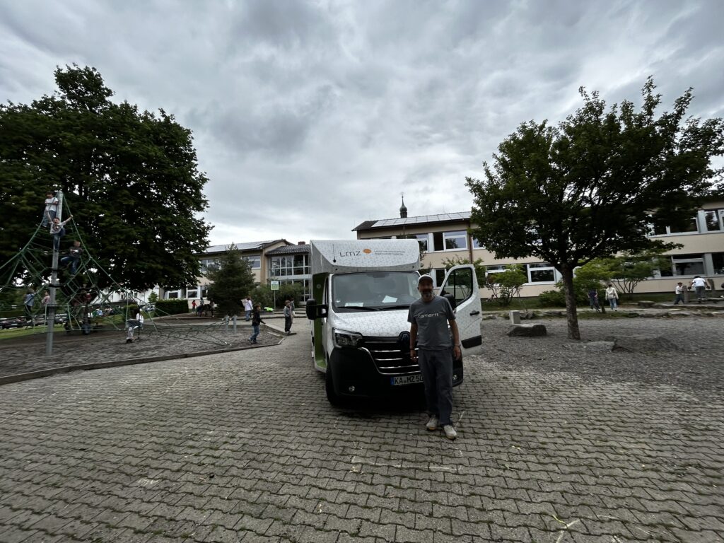 Ein weißer Transporter mit LMZ-Beschriftung steht auf dem gepflasterten Schulhof der Peter-Thumb-GMS Hilzingen. Davor steht eine Person mit Cap und grauem T-Shirt. Im Hintergrund sind Bäume, ein Klettergerüst mit Kindern und das Schulgebäude unter bewölktem Himmel zu sehen.