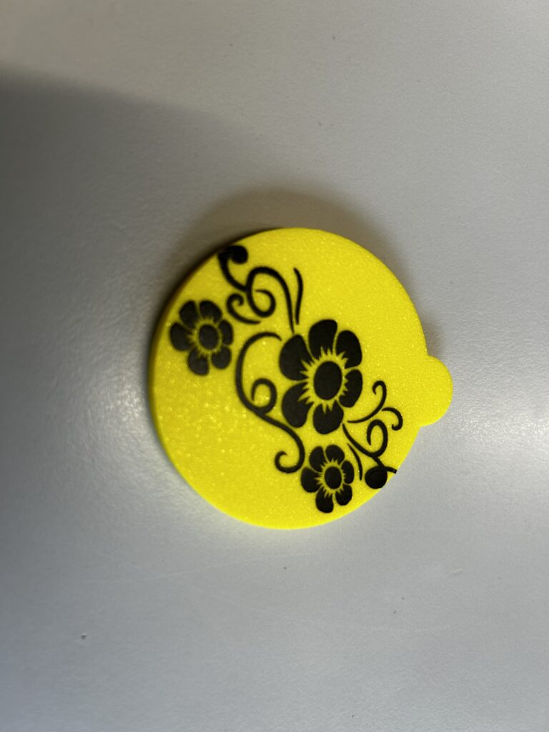 Ein runder Dosendeckel aus gelbem Kunststoff mit schwarz gelastertem Blumenmotiv – geschwungene Ranken und drei Blüten – liegt auf einer grauen Fläche.