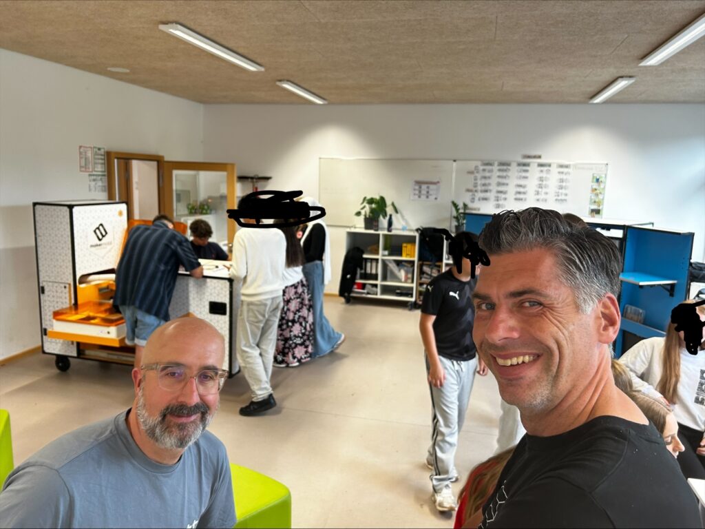 Selfie zweier erwachsener Männer im Vordergrund, beide lächeln in die Kamera. Im Hintergrund arbeiten mehrere Jugendliche an einem Lasercutter-Modul des Makermobils in einem hellen Klassenraum. Die Gesichter der Jugendlichen sind unkenntlich gemacht.