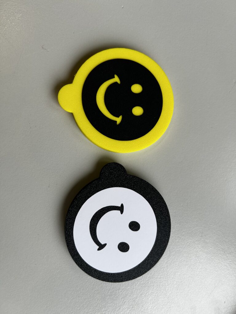 Zwei runde gelaserte Dosendeckel mit Smiley-Motiv liegen auf einer grauen Fläche. Der obere Deckel ist gelb mit schwarzem Hintergrund und gelbem Smiley, der untere ist schwarz mit weißem Hintergrund und schwarzem Smiley.