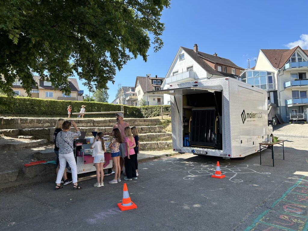 Makermobil auf Schulhof in Dingelsdorf, Kinder und Erwachsene am Grafik-Modul, blauer Himmel.