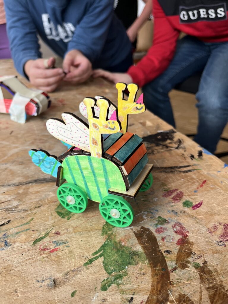 Ein Auto aus Holz in Grün und Gelb. Das GElb ist eine Giraffe, di eaus dem Auto schaut.