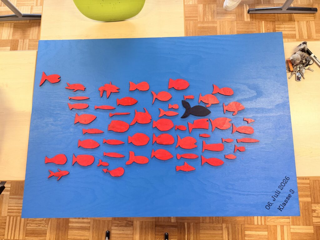 Blaue Holzplatte mit roten Holzfischen und einem schwarzen Fisch in der Mitte – nachgebaut aus der Geschichte „Swimmy", Klasse 3.