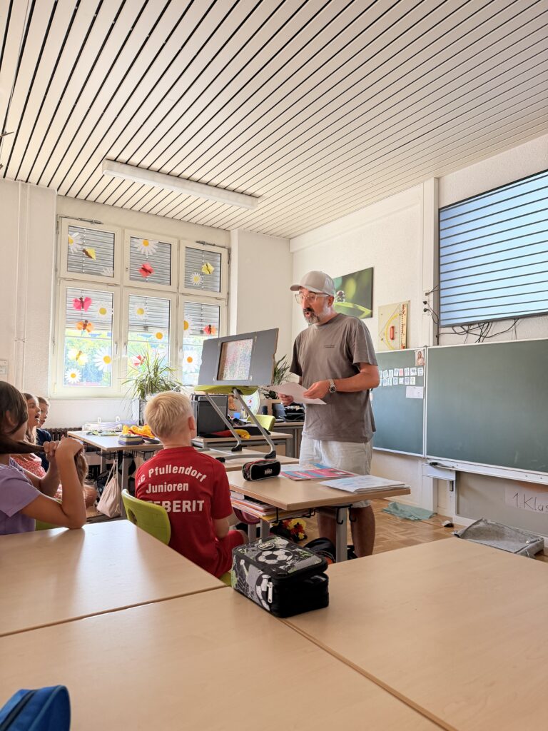 Ein Mann mit Cap und grauem T-Shirt steht vorne im Klassenzimmer und hält ein Blatt Papier in der Hand. Er spricht zu einer Gruppe von Grundschulkindern, die an ihren Tischen sitzen und zuhören. Im Hintergrund sind eine Tafel, ein Monitor und bunte Fensterdekorationen mit Blumen und Schmetterlingen zu sehen.