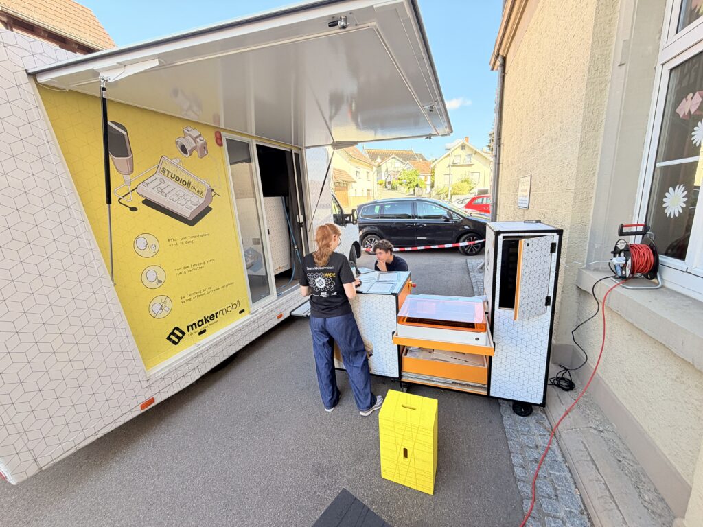 Das geöffnete Makermobil steht von innen sichtbar an einer Hauswand. Die gelbe Innenwand zeigt die Studio-Illustration mit dem Schriftzug „Studio on Air". Zwei Teammitglieder in schwarzen Makermobil-T-Shirts stehen vor dem ausgefahrenen Lasercutter-Modul in Orange-Weiß und bereiten den Einsatz vor. Auf dem Boden liegt ein gelber Würfel als Tritthilfe, seitlich ist eine rote Kabeltrommel zu sehen.