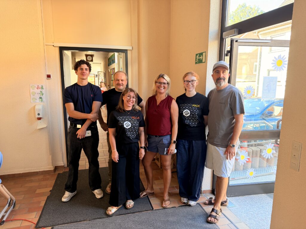 Sechs Personen stehen lächelnd im Eingangsbereich der Grundschule Zizenhausen. Drei von ihnen tragen schwarze Makermobil-T-Shirts mit den aufgedruckten Namen „Helga" und „Madie". Im Hintergrund ist ein Schulflur zu sehen, durch die Glastür rechts sind Gänseblümchen-Dekorationen erkennbar.